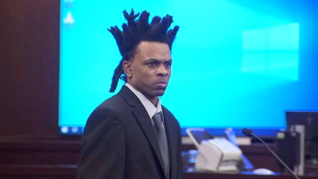 Ronnie O' Neal III wearing a suit and tie, with his distinctive tall, standing dreadlocks, appears serious and focused, standing in a courtroom.