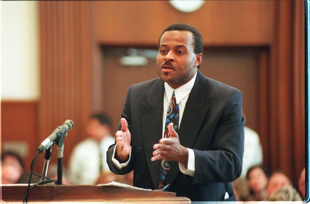 Colin Ferguson, wearing a suit, passionately speaking in a courtroom, gesturing with his hands, while seated individuals listen attentively.