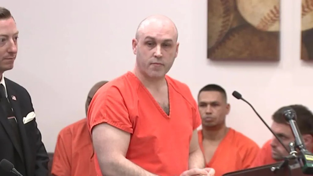 Trevor Summers, wearing an orange prison jumpsuit, stands at a podium during a court appearance, with a lawyer beside him.