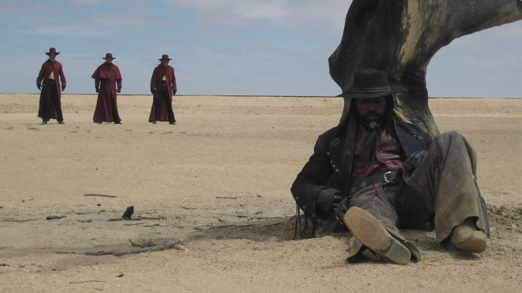 A scene from "Gallowwalkers". Aman (Snipes) sits on the ground in a barren landscape, with three figures in red cloaks and hats approaching in the background.