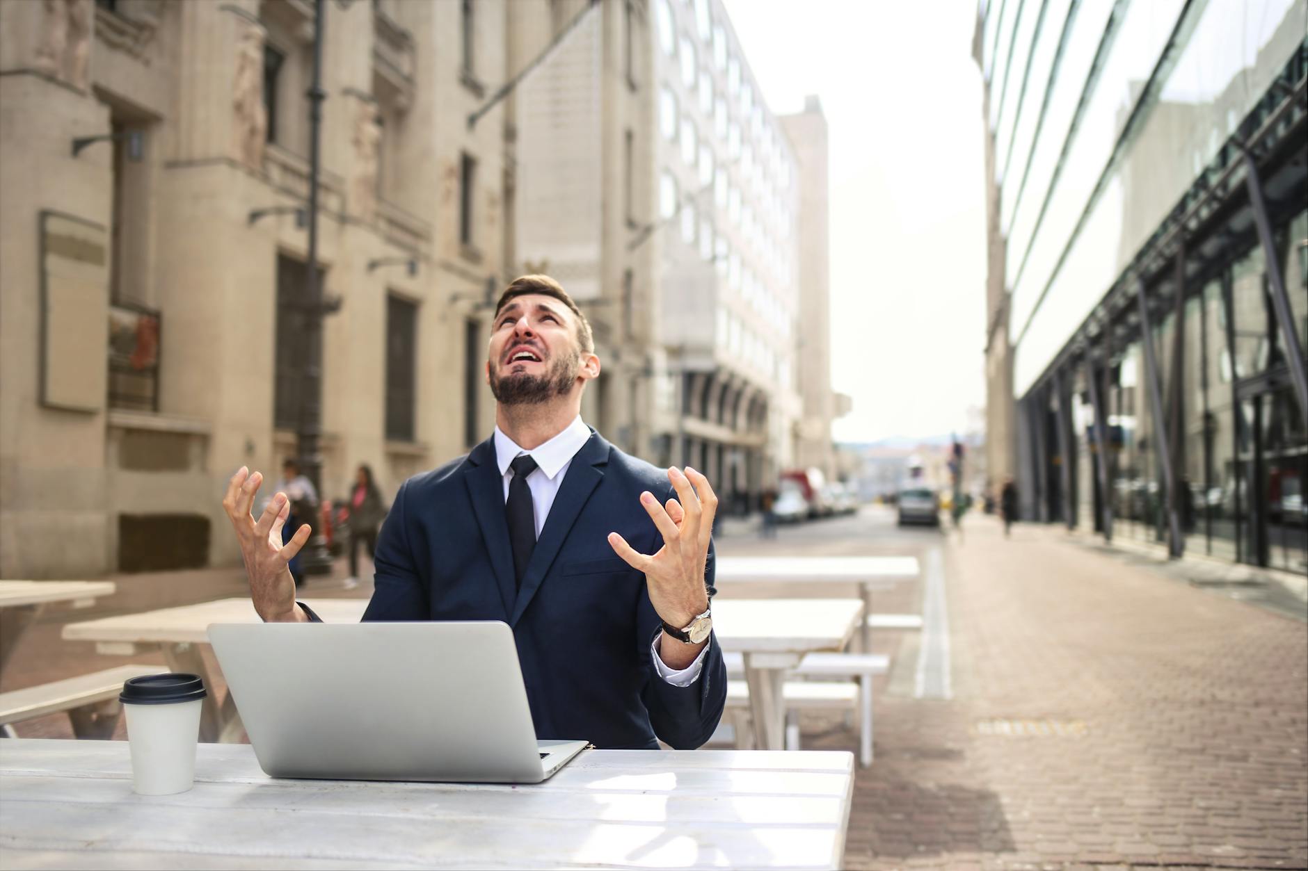 A frustrated businessman in a suit raises his hands in despair, sitting at an outdoor workspace with a laptop and coffee cup.