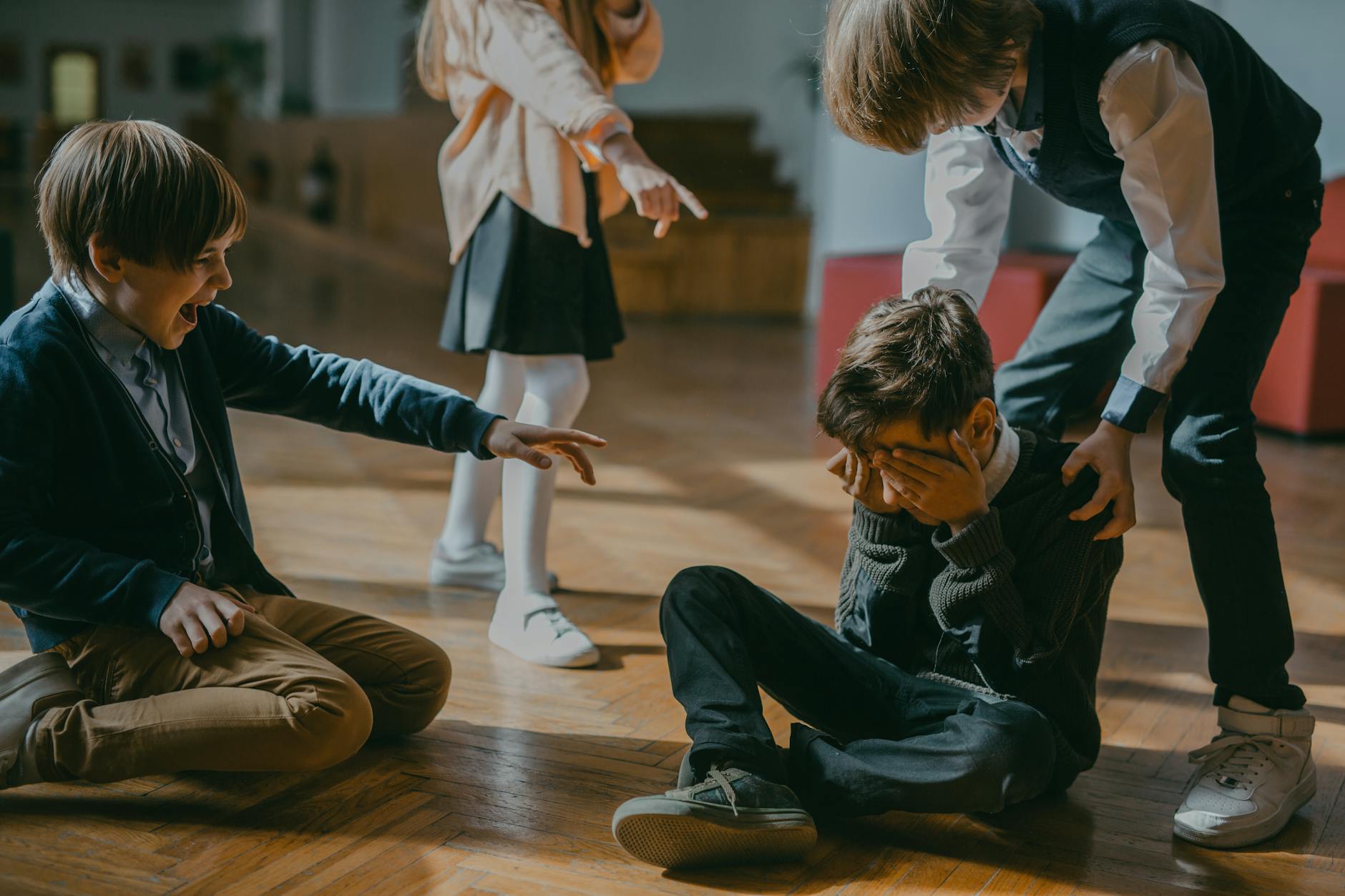 A group of four children in a school setting, with two boys laughing and pointing at a boy sitting on the floor, who has his face covered with his hands, appearing distressed. A girl in the background also points at him.