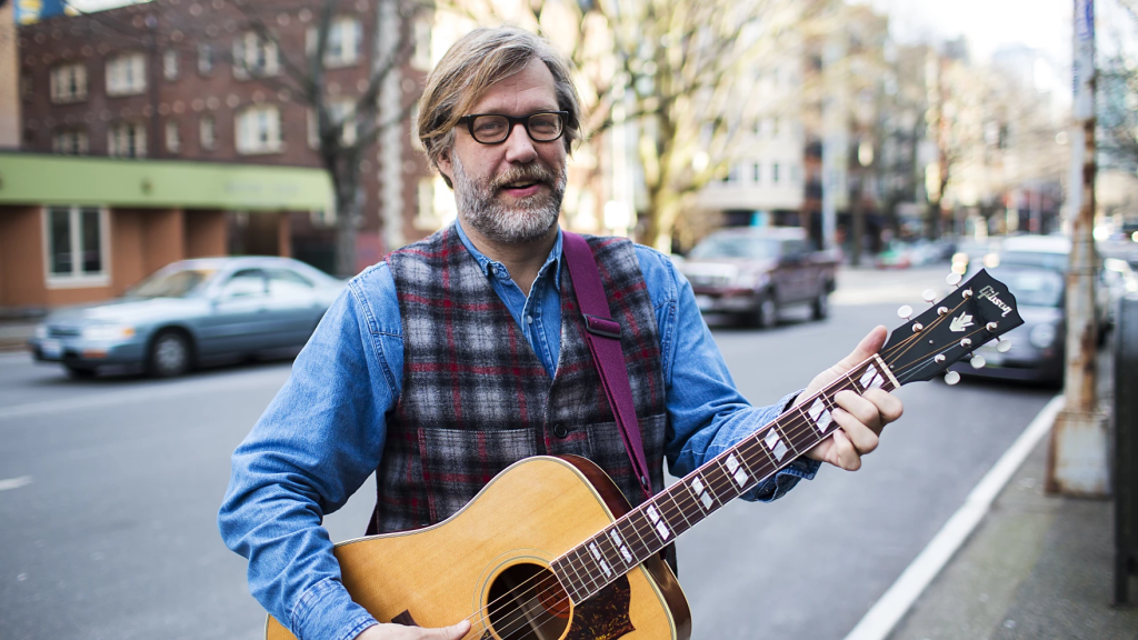 John Roderick, standing on a sidewalk, holding a guitar