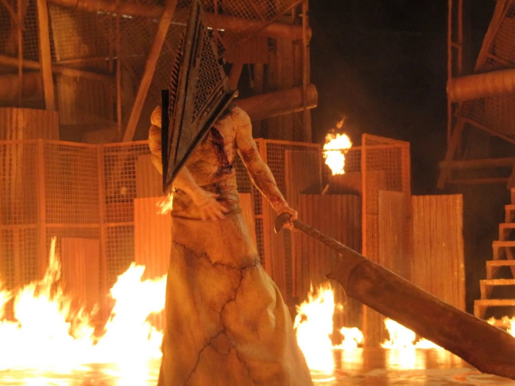 Screenshot from Silent Hill: Revelation. Pyramid Head stands before a wall of fire, holding his Great Knife to his side.
