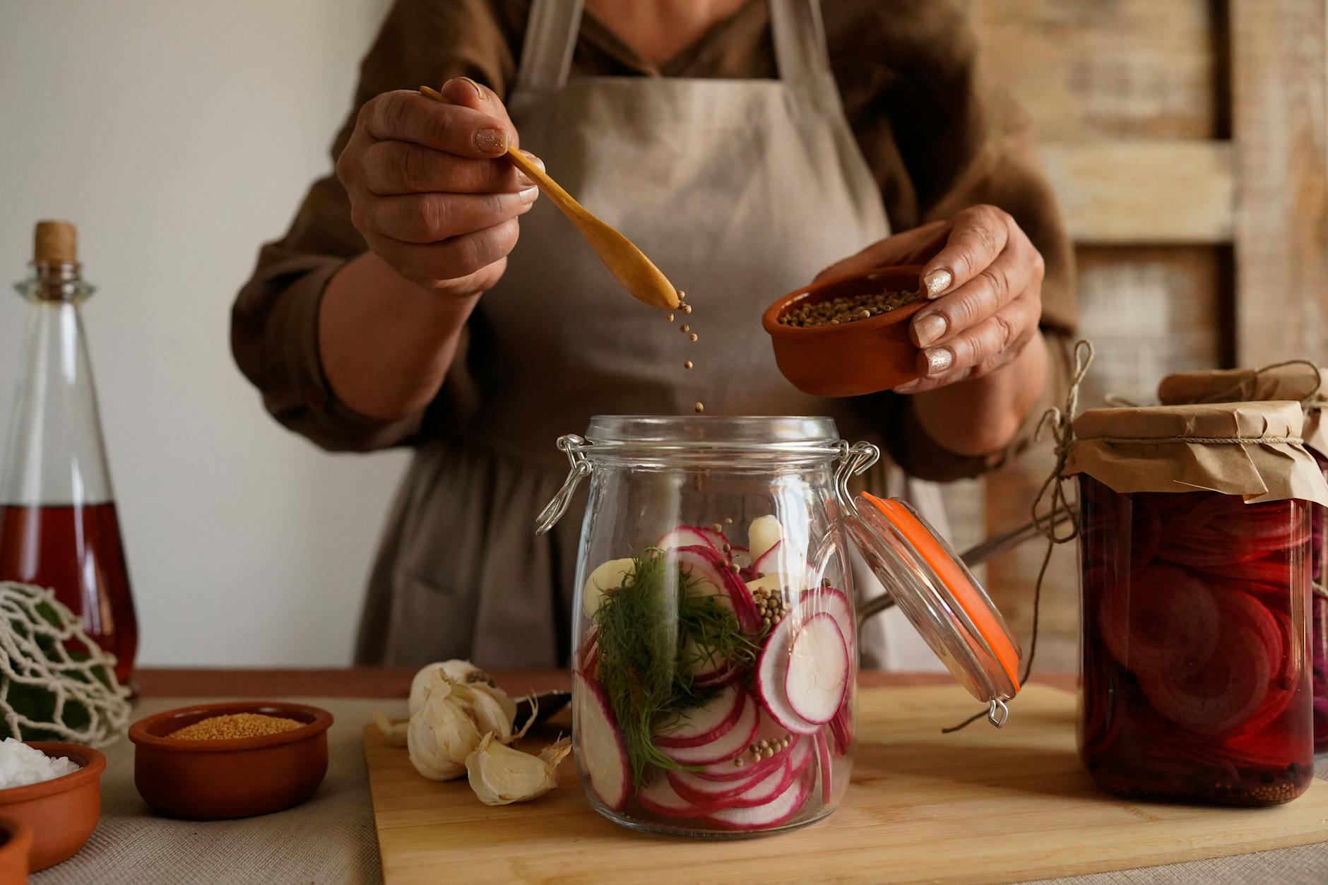 A person adding mustard seeds to a jar filled with sliced radishes and herbs, with garlic and other ingredients visible on a wooden cutting board.